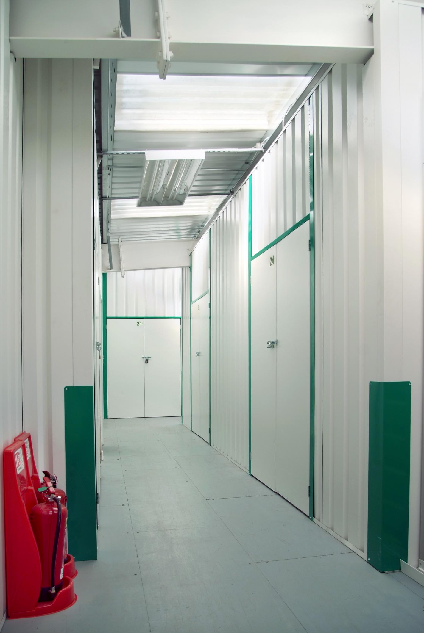 Hallway with metal storage unit doors and labeled rooms. A fire extinguisher and fire hose are mounted on the left wall.