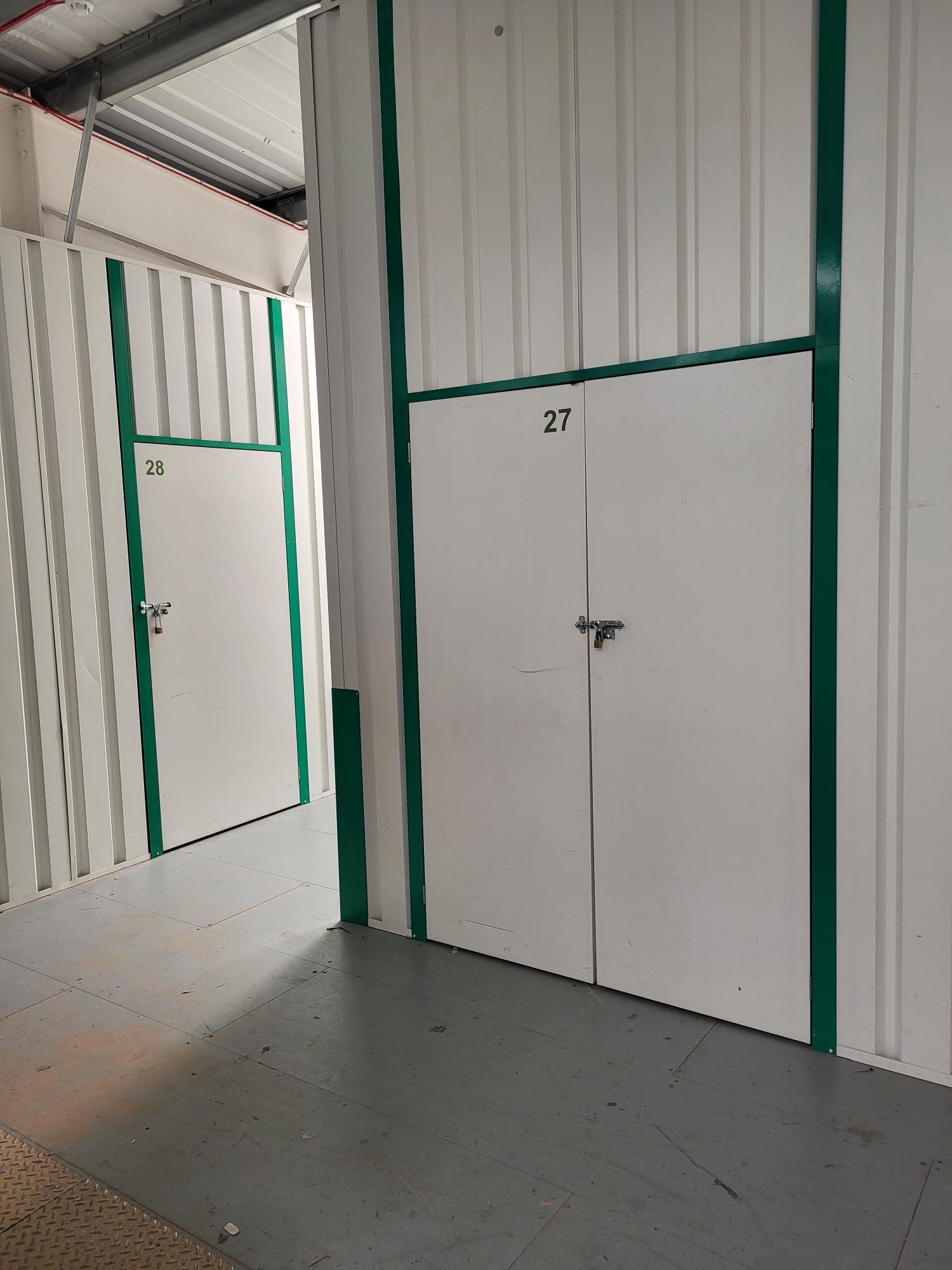 Two closed storage unit doors, numbered 27 and 28, with padlocks, inside a building with light gray flooring and white walls featuring green vertical stripes.