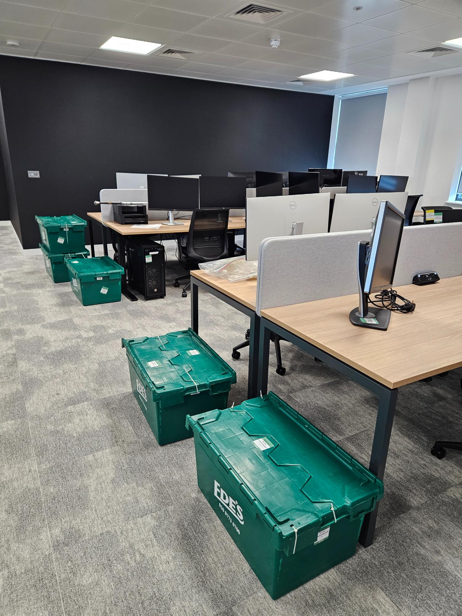 An office with desks, computer monitors, and several green plastic crates labeled "EDES" placed on the floor.