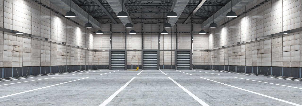 An empty industrial warehouse with multiple closed roller doors, grey concrete floor, and overhead lighting. White lines mark parking or storage areas on the floor. A yellow caution sign is visible.