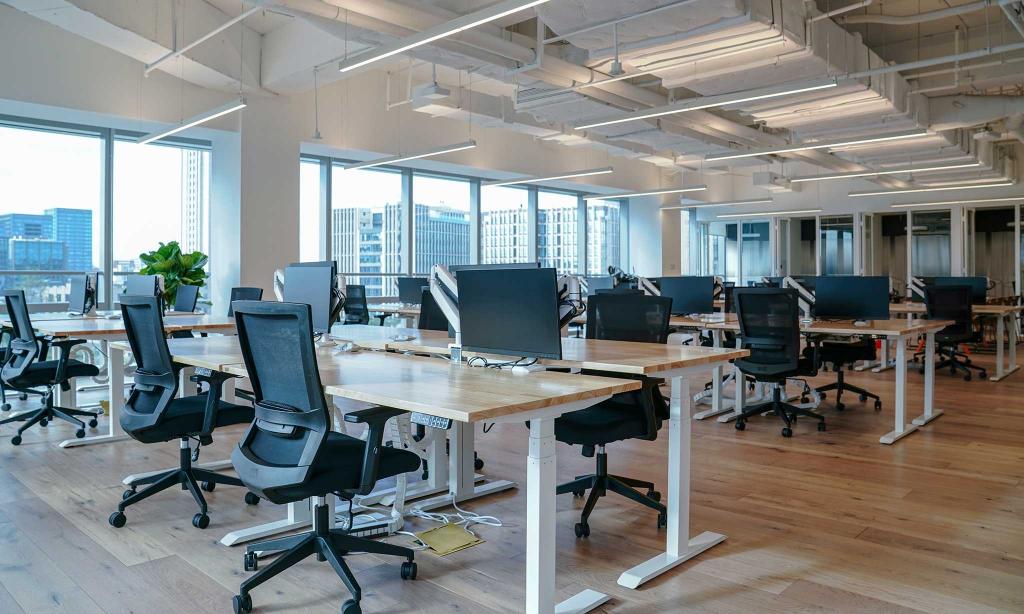 A modern, open-plan office space with multiple workstations featuring computers and black chairs, set against large windows overlooking city buildings.