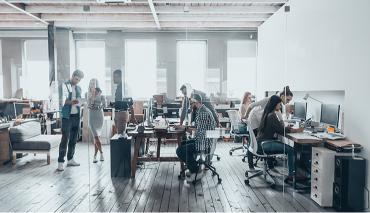 A modern office with an open-plan design where people are working at their desks. Some individuals are standing and conversing while others are engaged with their computers.