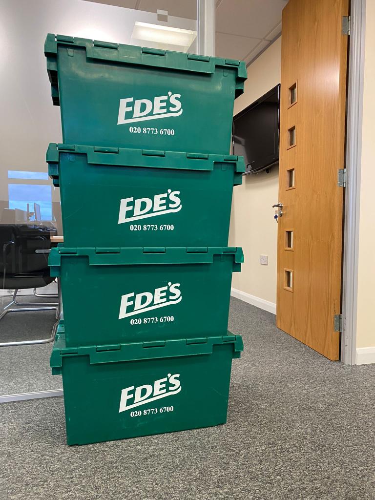 Four green plastic storage containers with the logo "EDE'S" and a phone number are stacked in an office setting near a wooden door and a glass partition.