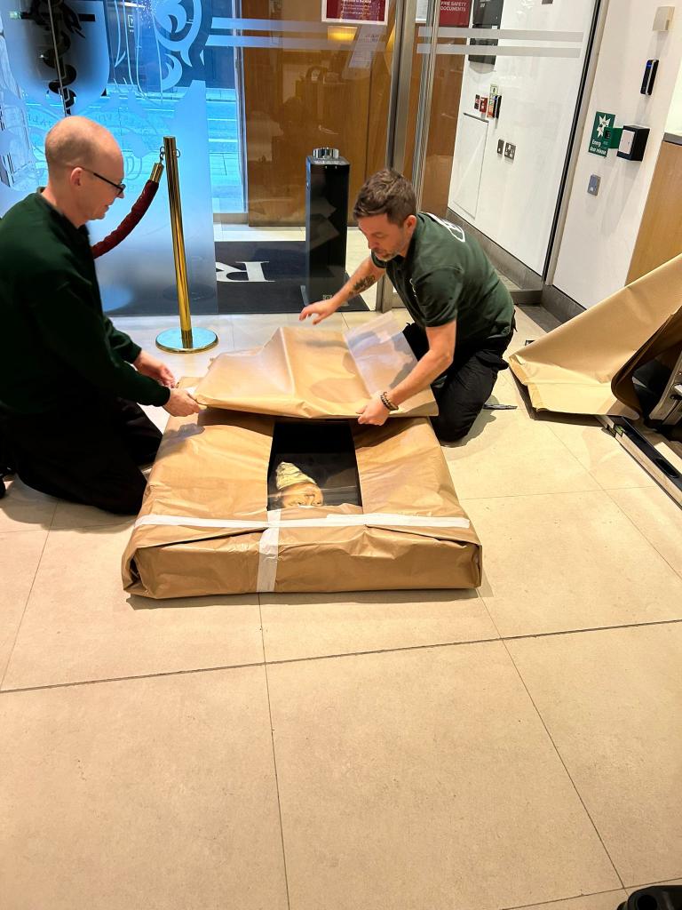 Two men are wrapping a large rectangular frame in brown paper, kneeling on the floor inside a building.