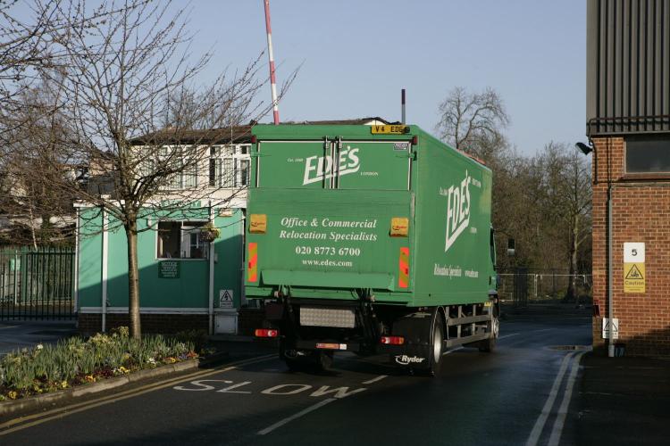 A green truck with "EDDS" branding is making a left turn, exiting a gated area with a barrier arm. The truck has a sign for "Office & Commercial Relocation Specialists". Trees and buildings are in the background.