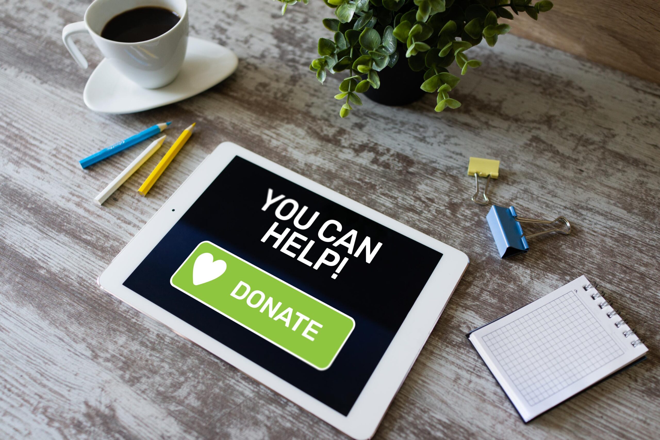 A tablet on a desk displays a "You Can Help!" Donate button. Nearby are a cup of coffee, a small plant, colored pencils, and a notepad with a binder clip.
