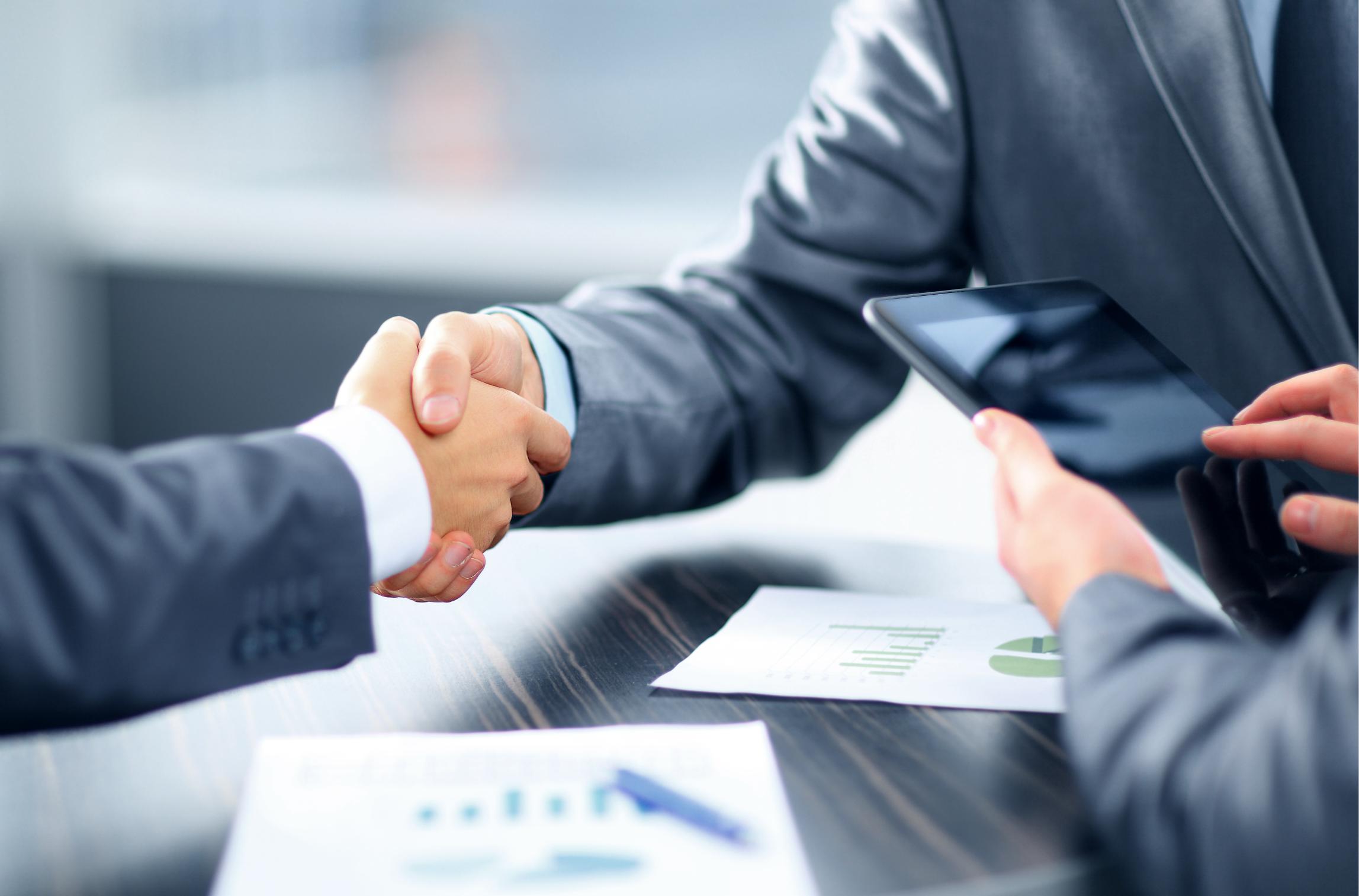 Two people in suits shake hands over a desk with business documents and a tablet, suggesting a business agreement or partnership.