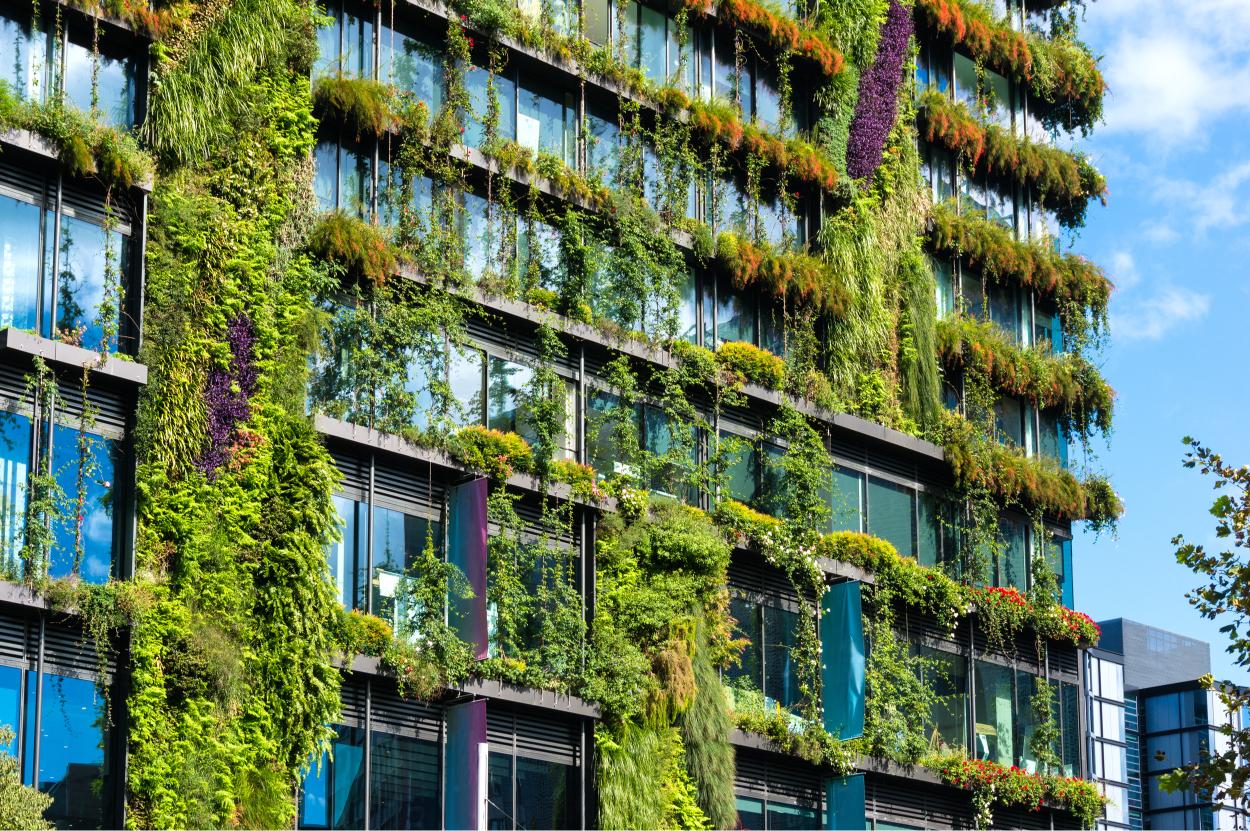 A multi-story building facade covered with lush vertical gardens, featuring various green plants and flowers growing along the entire exterior, set against a partly cloudy sky.