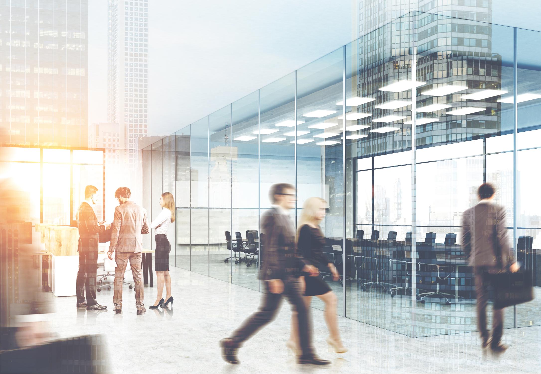 People walk and converse in a modern office space with glass walls, overlooking a city skyline.